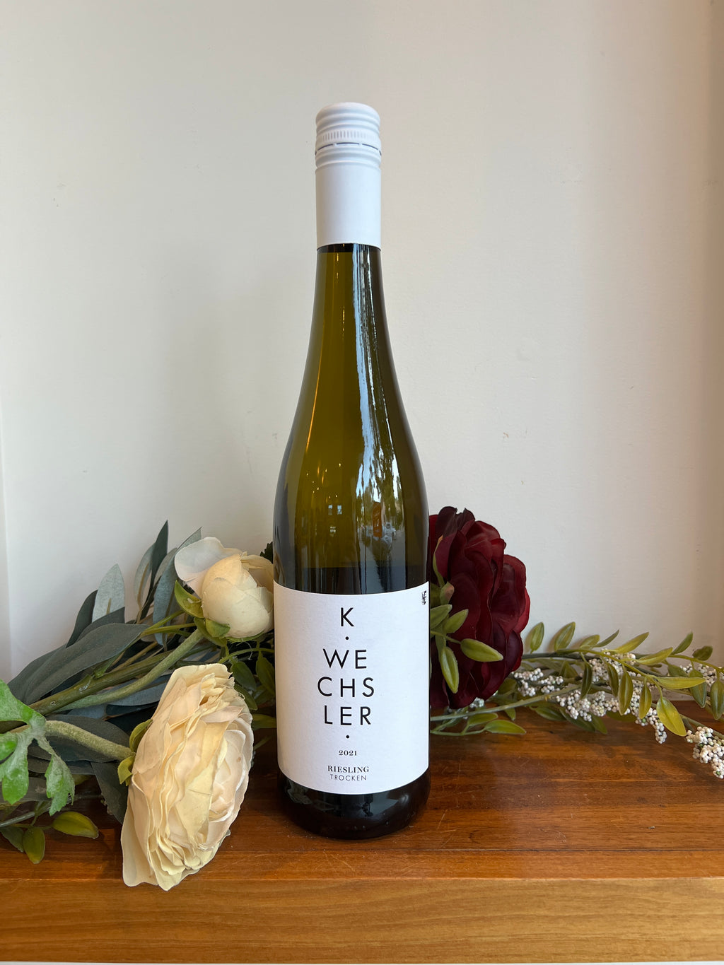 A bottle of Wechsler Dry Riesling wine positioned on a wooden surface with a floral arrangement and a decorative label.