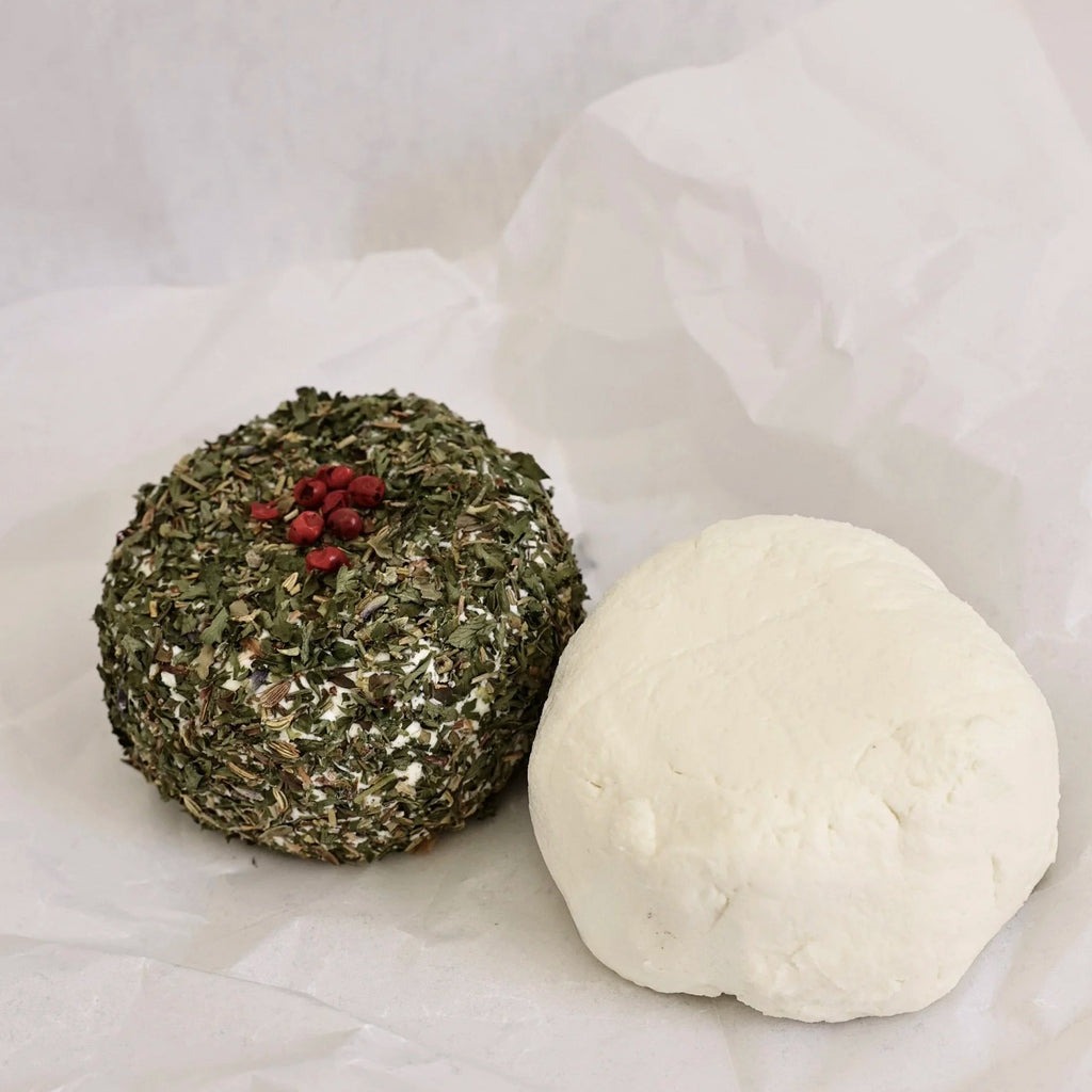 Two spherical cheeses, one green that is rolled in herbs and then added pink peppercorns atop, the other white, on a light gray/white background.