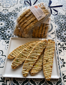 3 bags of 5 slices of pistachio biscotti, with 5 pieces laid out on a plate with slight white chocolate drizzle