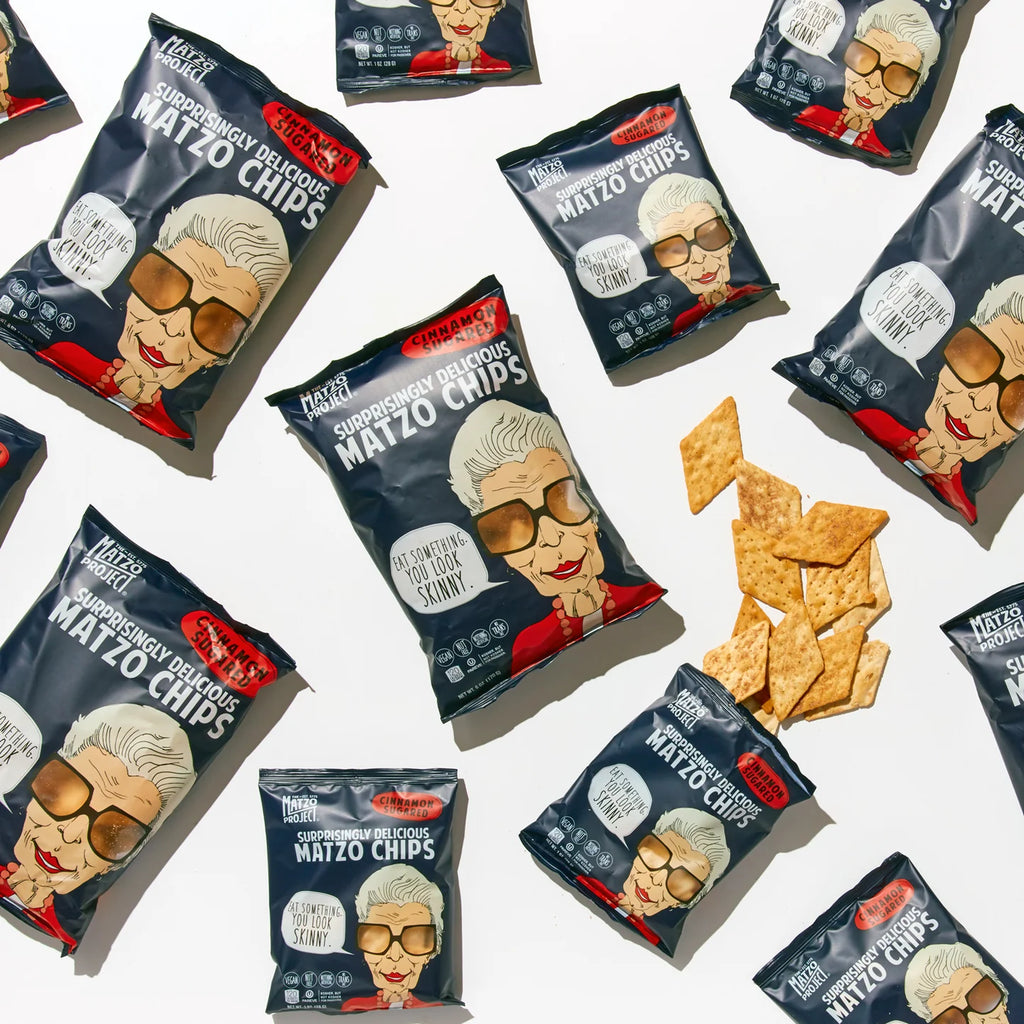 Multiple bags of Matzo Project Matzo Chips are arranged on a white surface, with several chips scattered unbagged at the center. Cinnamon Sugar Flavor