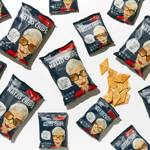 Multiple bags of Matzo Project Matzo Chips are arranged on a white surface, with several chips scattered unbagged at the center. Cinnamon Sugar Flavor