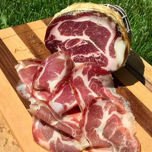 a chunk of Alle-Pia Coppa, with slices in front of it, on a wooden board outside on the grass