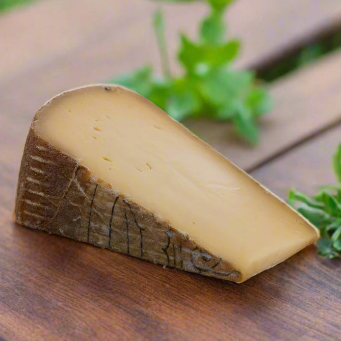Wedge of cheese, yellowy paste, no holes, with a dark brown rind, on wooden table with green leaves in background