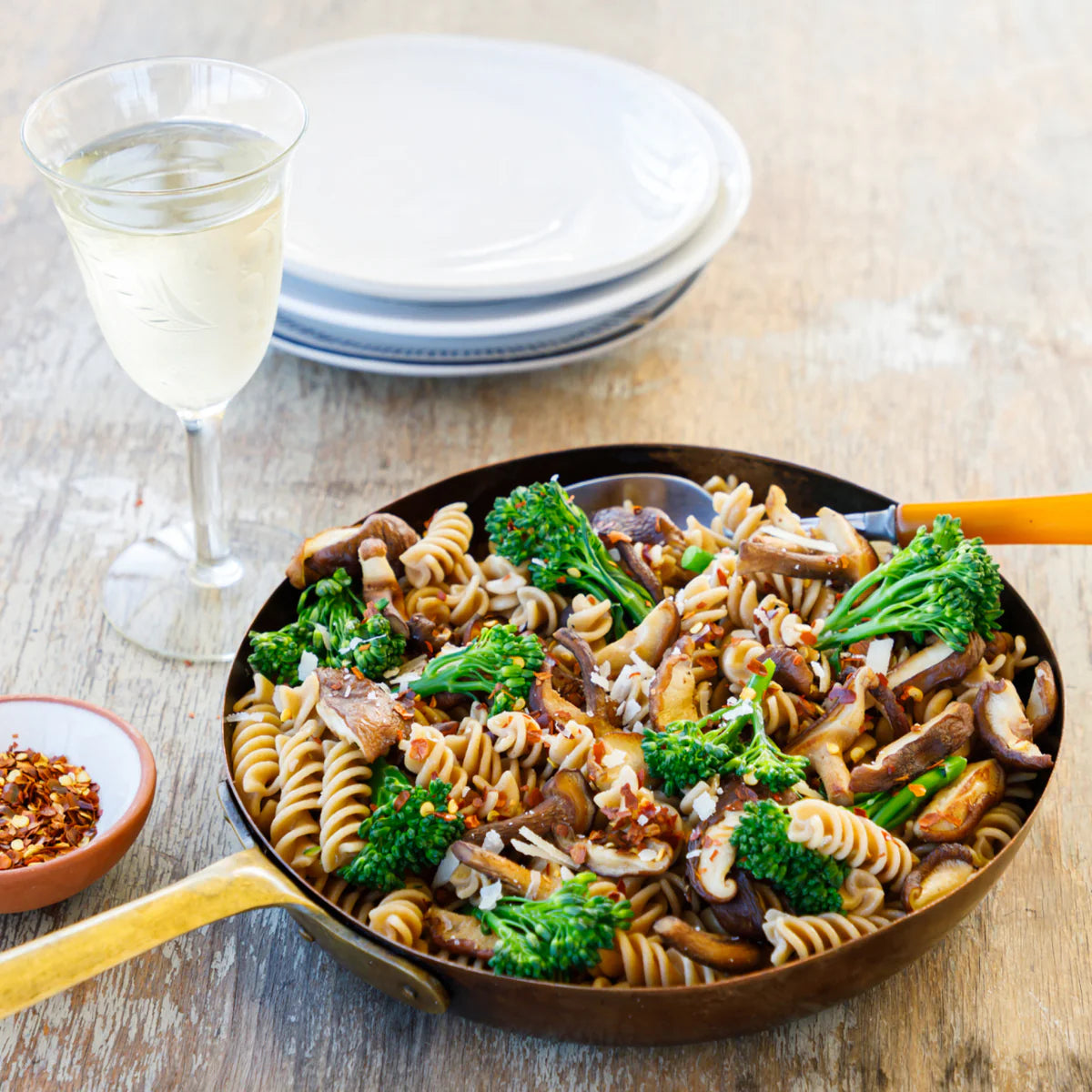 Pasta dish with broccoli and mushrooms in a pan on a wooden table with a glass of white wine.
