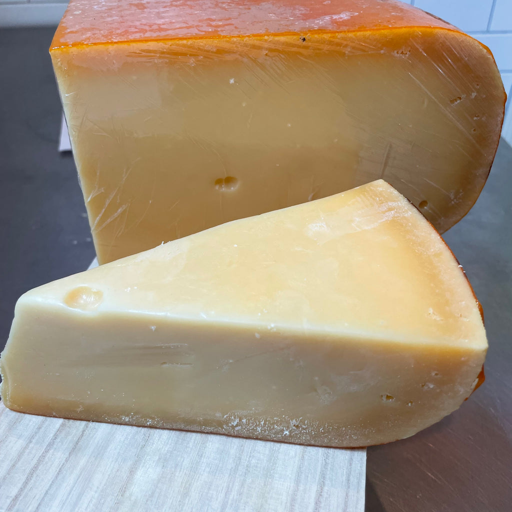 wedge of cheese with a pale golden paste, bright orange rind, a small amount of holes in the cheese, with a larger 1/4 wheel behind it on a pine wooden surface